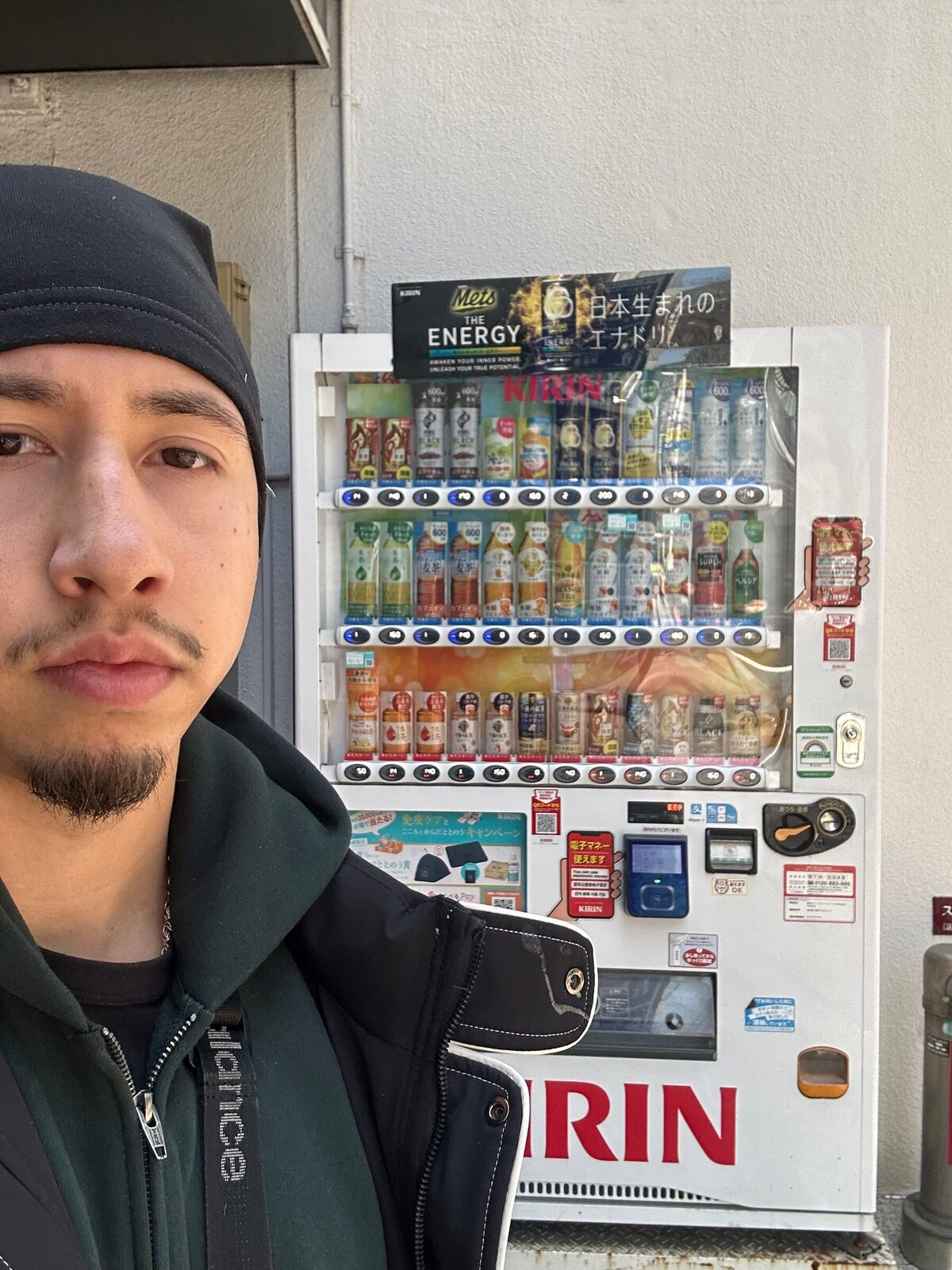 Eric står framför en dryckesautomat på en gata i Japan. Han bär mörk mössa och jacka, och bakom honom lyser automatens hyllor med flaskor och burkar upp mot en vit husvägg.