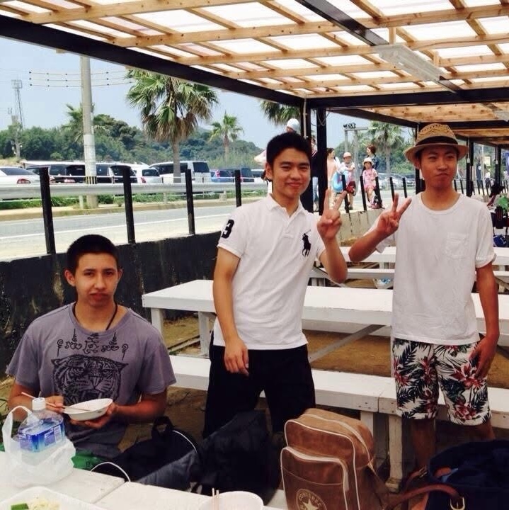 Tre unga män vid ett bord under ett soltak vid stranden i Fukuoka en solig sommardag 2014. En sitter vid bordet med en skål och ätpinnar i handen, två står bredvid och gör v-tecken mot kameran, och bakom dem syns bänkbord, palmer och en väg längs havet.