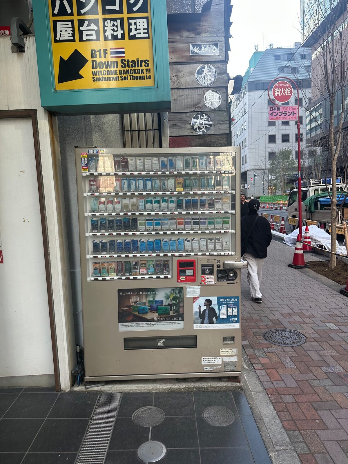 En beige cigarettautomat inbyggd i en husvägg på en gata i Tokyo. Bakom glaset syns rader av cigarettpaket, och under dem en betalpanel med myntinkast och kortläsare. En fotgängare passerar på trottoaren.